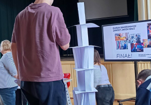 Uczeń podczas jednej z konkurencji konkursowych. Na stole widać wieżę z papieru, obok stoi uczeń.