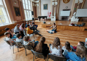 Uczniowie siedzą na krzesełkach w sali. Przed nimi widać osobę prowadzącą zajęcia, klęczącą przed manekinem.