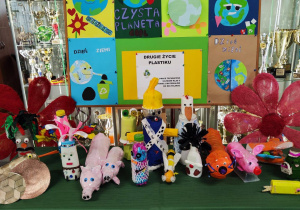 Na stoliku przykrytym zielonym suknem stoją prace - zwierzątka, stworki i pojazdy wykonane z plastiku. Z tyłu stoi tablica z powieszonymi plakatami z okazji Dnia Ziemi.