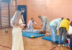 W sali gimnastycznej uczniowie stoją przy materacach, nachylają się nad osobami leżącymi na materacu.