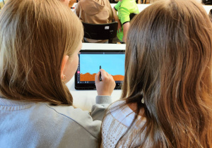 Na stole stoi tablet, na który patrzą dwie dziewczynki.