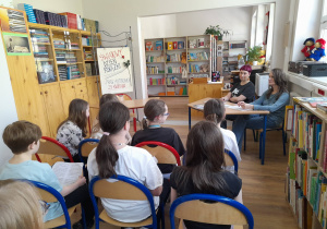 W bibliotece szkolnej na krzesełkach siedzą uczniowie. W głębi kadru widać jury konkursowe.