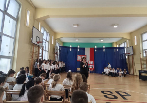 W sali gimnastycznej uczniowie ubrani w stroje galowe siedzą na krzesełkach.
