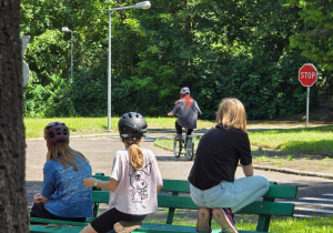 Uczniowie siedzący na ławce obserwują koleżankę jeżdżącą na rowerze po uliczkach motodromu.