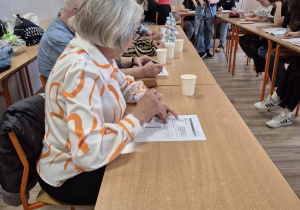 W pomieszczeniu w rzędem ustawionych ławkach siedzą seniorzy oraz uczniowie.