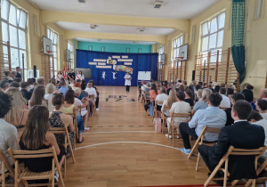 W sali gimnastycznej na krzesłach siedzą uczniowie. W głębi na granatowym suknie wisi dekoracja.