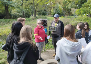 W parku na ścieżce stoi grupa uczniów. Słuchają opowieści prowadzącego widocznego w głębi kadru.