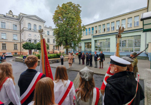 Na dziedzińcu szkolnym stoi poczet sztandarowy. W głębi kadru widać uczestników uroczystości szkolnych.