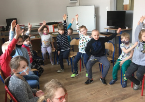 Uczniowie klasy 1a siedzą w bibliotece na krzesełkach; niektóre dzieci podnoszą ręce w górę zgłaszając chęć odpowiedzi