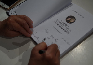 na zdjęciu widać dwie ręce, w prawej jest długopis, ręce oparte są o otwartą książkę, na której jest autograf