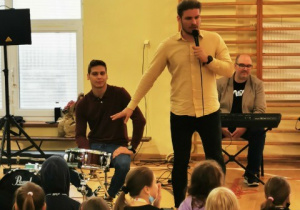 uczniowie siedzą na sali gimnastycznej, jeden z muzyków siedzi przy perkusji, drugi przy keyboardzie, prowadzący opowiada o perkusji