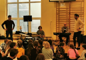 uczniowie siedzą na sali gimnastycznej, przy perkusji jeden z uczniów, prowadzący i perkusista przyglądają się co robi uczeń, na dalszym planie przy keyboardzie siedzi muzyk