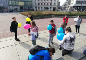 na zdjęciu grupka naszych uczniów stoi na placu Wolności i nadmuchuje kolorowe balony