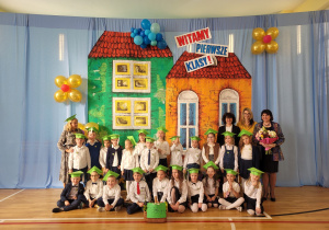 zdjęcie grupowe uczniów klasy 1b wraz z wychowawcą oraz trzema paniami dyrektor, na sali gimnastycznej, w tle dekoracja, na którą składają się dwa domki i balony