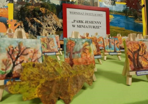 na stole przykrytym zielonym suknem stoją prace dzieci, są to miniaturowe obrazki na miniaturowych sztalugach przedstawiające jesienny krajobraz w parku