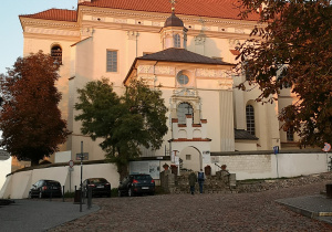 Zdjęcie przedstawia renesansowy Kościół Farny w Kazimierzu Dolnym. Kościół otoczony położony jest na wzgórzu i otoczony murem. Wejście na dziedziniec kościoła wytyczają schody. Na zdjęciu widać ludzi, którzy wchodzą do kościoła.