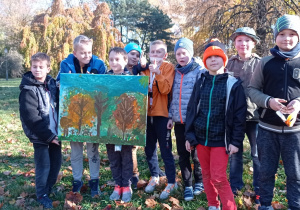 Zdjęcie przedstawia grupę chłopców z klasy 3a, którzy wykonali jesienny pejzaż na płótnie. Prezentują swój obraz w parku, za nimi widoczne są drzewa w jesiennych barwach.