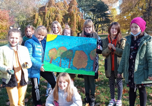 Na zdjęciu uczennice z klasy 3a trzymają w dłoniach obraz, który wykonały z wykorzystaniem farb plakatowych. Obraz przedstawia pejzaż jesienny. W tle za dziewczynkami widać drzewa w jesiennych barwach. Dziewczynki ubrane są w kurtki, są uśmiechnięte.