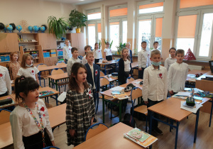 uczniowie w klasie ubrani w stroje galowe, stoją na baczność, śpiewają hymn