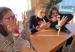 Trzy dziewczynki siedzą razem w ławce, pokazują ozdoby papierowe w barwach polskich i ukraińskich