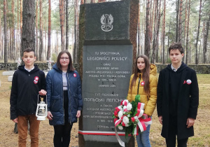 Czworo uczniów stoi obok pomnika w Polskim Lasku. Pomnik upamiętnia polskich legionistów. Chłopiec po lewej stronie trzyma znicz, dziewczynka po prawej trzyma wiązankę