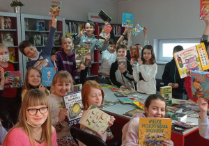 Zdjęcie przedstawia uczniów z klasy 3a podczas warsztatów w Bibliotece Miejskiej. Uczniowie siedzą na krzesłach trzymając w dłoniach kolorowe książki. Uczniowie są w dobrych humorach, uśmiechają się.