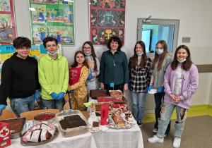grupa uczniów szkolnego szkoła PCK wraz z panią dyrektor stoją za stołem, na którym są ciasta przygotowane na kiermasz