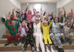 uczniowie wraz z nauczycielkami siedzą na schodach przed salą gimnastyczną, są ubrani w piżamy