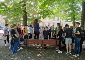 grupa uczniów klas siódmych stoi w parku słuchając nauczyciela, który opowiada historę przemysłowej Łodzi