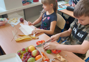 dwoje dzieci siedzących w ławce przygotowuje warzywno-owocowy szaszłyk