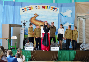 scena z przedstawienia - dziewczynka ubrana w barwy polskie owinięta czarną chustą stoi otoczona grupą uczniów ubranych w stroje żołnierskie