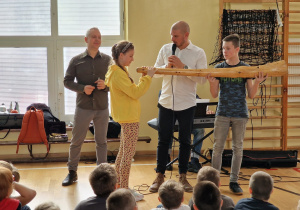 uczniowie siedzą na podłodze sali gimnastycznej, przyglądają się jak dwoje uczniów próbuje grać na drewnianym instrumencie