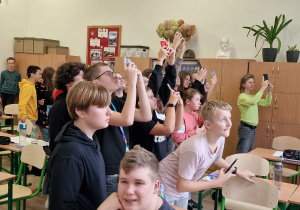 uczniowie stoją w klasie przy oknie, trzymają w rękach telefony i obserwują zjawiskok częściowego zaćmienia słońca