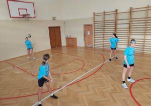 na sali gimnastycznej stoją cztery dziewczynki ubrane w stroje siatkarskie, jedna z nich serwuje piłkę