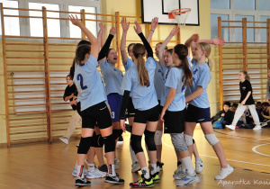na sali gimnastycznej drużyna dziewcząt stoi zgromadzona w kółko, z rękami podniesionymi do góry w geście zwycięstwa
