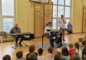 Na sali gimnastycznej muzycy demonstrują możliwości perkusji elektronicznej. Jeden z muzyków gra na keyboardzie. W tle widać również mężczyznę prowadzącego koncert. Przodem do muzyków na podłodze siedzą uczniowie.