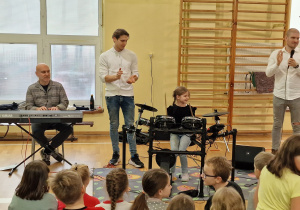 Uczennica siedzi przy perkusji elektronicznej, trzyma w dłoniach pałki perkusyjne. Na keyboardzie gra jeden z muzyków. Drugi wraz z prowadzącym i widownią złożoną z uczniów przysłuchują się grze uczennicy.