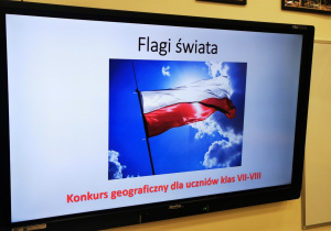 Na zdjęciu widoczny jest telewizor z wyświetloną prezentacją multimedialną. Na ekranie widoczna jest flaga Polski i napis - Flagi świata, konkurs dla uczniów klas VII-VIII.