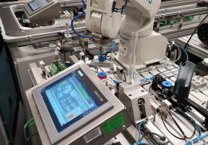 Zdjęcie przedstawia zautomatyzowaną linię produkcyjną, na której pracuje robot, który składa plastikowe elementy oraz monitor, na którym widać schemat linii produkcyjnej.