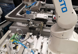 Zdjęcie przedstawia zautomatyzowaną linię produkcyjną, na której ramię robota składa plastikowe elementy.