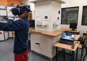Zdjęcie przedstawia ucznia, który ma na sobie gogle 3 D i porusza się po wirtualnym świecie. Z boku stoi laptop, na którym widać środowisko, w którym porusza się uczeń.
