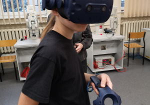 Zdjęcie przedstawia ucznia, który ma na sobie gogle 3 D i porusza się po wirtualnym świecie. Uczeń w rękach joysticki, dzięki którym steruje i porusza się po wirtualnym świecie.