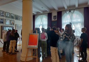 Uczniowie stoją w sali. Widać kolumnę oraz zdjęcia wiszące na ścianie po lewej stronie kadru