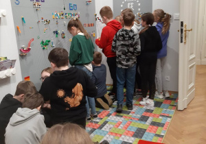 Uczniowie oglądają wystawę klocków Lego. Na ścianie umieszczone są różne figurki oraz drobne elementy zbudowane z klocków. Na podłodze leży dywan z grafiką w klocki.