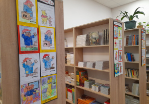 Na zdjęciu widać regały z książkami w bibliotece szkolnej. Na szczytach tych regałów wywieszone są prace uczniów. Przedstawiają one pokolorowane misie, między innymi są to misie z różnych bajek.