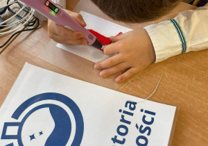 Ręce ucznia trzymające długopis 3d oraz praca ucznia - flaga Polski. Na stole leży również kartka z logo "Laboratoria Przyszłości"