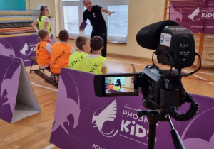 Na pierwszym planie ustawiona kamera nagrywa osobę prowadzącą zajęcia sportowe, które tłumaczy dzieciom przebieg konkurencji.