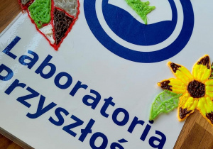Na stole leżą grzybek, listek, kwiatek wykonane długopisem 3d, pod spodem znajduje się logo programu Laboratoria Przyszłości