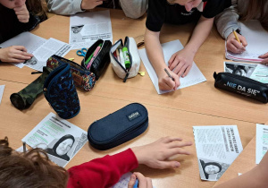 Na zdjęciu widać dwie złączone ławki. Na ławkach leżą kartki, na których uczniowie piszą listy. Widać ich ręce z długopisami, piórniki i kartki informacyjne o maratonie pisania listów.