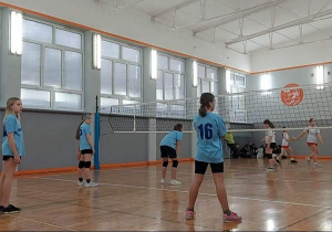 Na sali gimnastycznej fragment meczu piłki siatkowej. Po obu stronach siatki stoją drużyny dziewczęce. Bliżej drużyna w niebieskich koszulkach. Dziewczynki stoją tyłem.